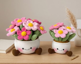 May include: Two crochet flower pots with smiling faces and brown feet sit on a wooden surface. The flowers are pink with yellow centers and green leaves. The pots are white.