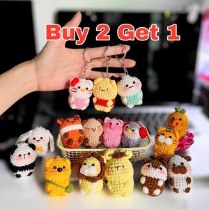 Puede incluir: Una colección de llaveros de animales de ganchillo coloridos, incluyendo un panda, un oso y un león. Los llaveros son de una variedad de colores y estilos. El texto "Buy 2 Get 1" se muestra en rojo.
