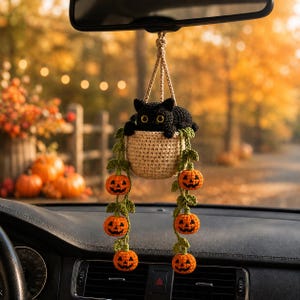 Peut inclure: Un ornement de voiture crocheté représentant un chat noir assis dans un panier beige. Deux vignes vertes avec de petites citrouilles d'Halloween orange pendent du panier. L'ensemble est suspendu par un cordon beige à un rétroviseur.