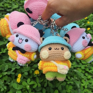 May include: A collection of handmade crochet keychains featuring cartoon characters. Each keychain has a unique design, including hats, outfits, and small bags. The keychains are in various colors, including pink, yellow, blue, and green.