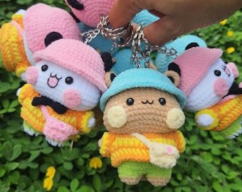 May include: A collection of crochet plush keychains featuring small characters wearing colorful bucket hats, sweaters, and crossbody bags. Each plush has black eyes, a stitched mouth, and is attached to a silver metal keyring for hanging.