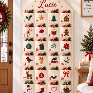 Peut inclure: Un calendrier de l'Avent mural en tissu crème en forme de maison, portant le prénom Lucie en haut. Il comporte vingt-quatre poches numérotées, chacune décorée d'icônes de Noël uniques telles qu'un bonhomme de neige, un renne et des cadeaux.