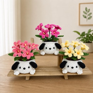 May include: Three crocheted potted plants featuring white puppy-shaped containers with black ears and paws. The left pot holds pink flowers, the center pot holds magenta flowers, and the right pot holds yellow flowers, all with green leaves.