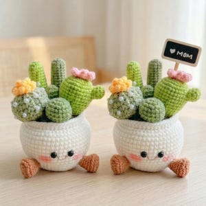 May include: Two handmade crochet cactus plants in white pots with smiling faces and small brown feet. The cacti are green with yellow and pink flower accents and pearl embellishments. One pot has a small sign that says "MOM".