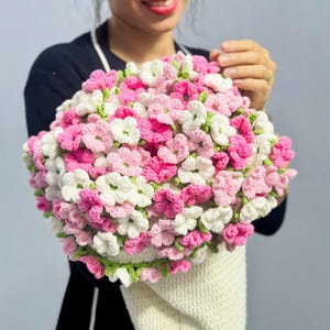 May include: A bouquet of crocheted flowers in shades of pink and white, with green accents, held in a cream-colored crocheted cone. The flowers are densely packed, creating a full, textured appearance.