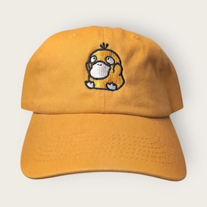 Puede incluir: Gorra de béisbol de color amarillo mostaza con un diseño de pato de dibujos animados bordado en la parte delantera. La gorra tiene una visera curva y una correa ajustable en la parte trasera. El pato es amarillo con cara blanca y contorno negro.