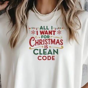 Pode incluir: Camiseta creme com a frase "ALL I WANT FOR CHRISTMAS IS CLEAN CODE" em vermelho, verde e branco. A camiseta tem mangas curtas e um corte folgado.
