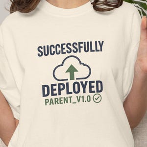 Op de afbeelding: Een crèmekleurig T-shirt met korte mouwen en de donkerblauwe tekst "SUCCESSFULLY DEPLOYED" boven een wolkenpictogram met een groene pijl die omhoog wijst. Onder de wolk staat de tekst "PARENT_V1.0" in het groen met een groen vinkje.