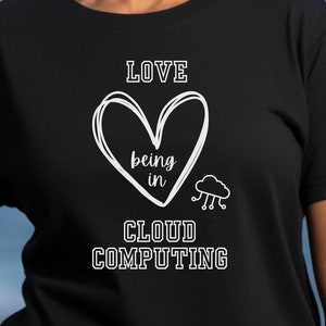 May include: Black t-shirt featuring the white text "LOVE being in CLOUD COMPUTING", a heart, and a cloud with rain. The shirt is worn by a person, with a blurred ocean backdrop.