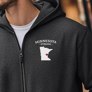 K&ouml;nnte beinhalten: Anthrazitfarbener Kapuzenpullover mit Rei&szlig;verschluss, der den Text "MINNESOTA STRONG" und eine wei&szlig;e Umrisslinie des Bundesstaates Minnesota mit einem roten Herzen zeigt. Der Hoodie hat eine Kapuze und einen Frontrei&szlig;verschluss.