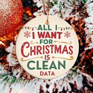 May include: A round Christmas ornament with the text "ALL I WANT FOR CHRISTMAS IS CLEAN DATA" in red, green, and gold. The ornament hangs on a snowy Christmas tree with a glittery orange ball.
