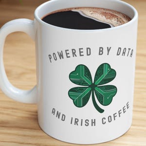 May include: White ceramic mug with a handle, filled with dark coffee. The mug features the text "POWERED BY DATA AND IRISH COFFEE" and a green four-leaf clover design. The mug is on a wooden surface.