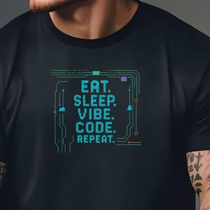 以下が含まれることがあります： 「EAT. SLEEP. VIBE. CODE. REPEAT.」の文字がピクセル化された基板風のデザインでプリントされた黒いTシャツ。文字は青と緑の色合いです。Tシャツはクルーネックです。