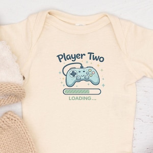 May include: A cream babygrow featuring the text "Player Two" and a video game controller graphic. The image also shows a knitted fox hat, matching nappy cover, brown baby boots, and a wooden rattle.