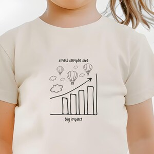 May include: A cream-colored t-shirt with a black graphic of hot air balloons, clouds, and a rising bar graph. The text "small sample size" and "big impact" are above and below the graphic. The person is wearing floral shorts.