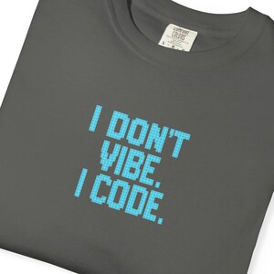 Puede incluir: Camiseta gris oscuro con el texto "I DON'T VIBE. I CODE." en fuente pixelada azul claro. La camiseta tiene cuello redondo y una pequeña etiqueta en la parte superior derecha. La camiseta está doblada.