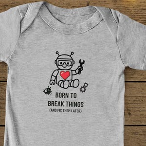 Op de afbeelding: Grijze babybody met een robotafbeelding met een sleutel en een hart. De tekst "BORN TO BREAK THINGS (AND FIX THEM LATER)" staat eronder. De afbeelding bevat een paar zwarte babyschoenen. Achtergrond van hout.