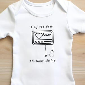 以下が含まれることがあります： 白いベビーロンパースに、心臓モニターの白黒のグラフィックと「tiny resident 24-hour shifts」のテキストがプリントされています。