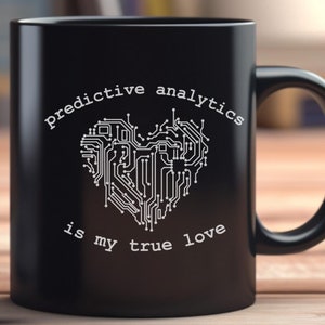 May include: Two black ceramic mugs with the text "predictive analytics is my true love" in white. The text is above a heart-shaped design made of circuit board lines. The mugs are on a wooden surface.