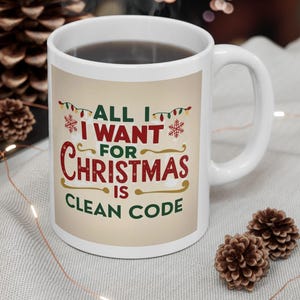 May include: White ceramic mug with the text "ALL I WANT FOR CHRISTMAS IS CLEAN CODE" in red, green, and gold. The mug is filled with a dark liquid and has a white handle. The mug is on a light grey surface with Christmas decorations.