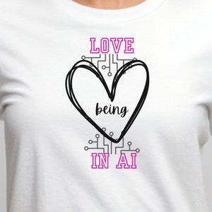 May include: White t-shirt featuring a black heart design and the words "LOVE being in AI" in pink. The heart is outlined with a black line and the text is in a sans-serif font. The shirt is made of a soft material.