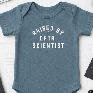 Könnte beinhalten: Ein kurzärmeliger Babybody in meliertem Blau mit dem weißen Schriftzug "RAISED BY A DATA SCIENTIST" auf der Brust. Das Kleidungsstück hat einen Rundhalsausschnitt, kurze Ärmel und einen Druckknopfverschluss im Schrittbereich.