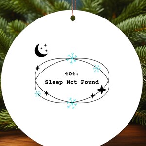 May include: White round Christmas ornament with the text "404: Sleep Not Found" in black. The ornament features a crescent moon, stars, and snowflakes. The ornament hangs from a brown string, with a Christmas tree and other ornaments in the background.