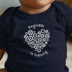 Könnte beinhalten: Marineblauer Baby-Body mit dem Text "engineer in training". Der Text ist weiß und zeigt ein herzförmiges Design aus Zahnrädern. Der Body hat kurze Ärmel.