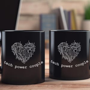 Könnte beinhalten: Zwei schwarze Kaffeetassen mit einem weißen Herzdesign aus Leiterplatten und den Worten "tech power couple". Die Tassen stehen auf einer Holzoberfläche, mit einem Laptop und Büchern im Hintergrund.