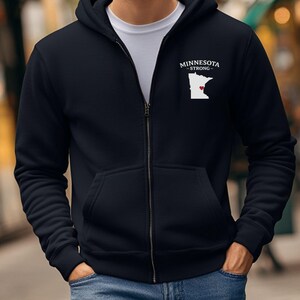 K&ouml;nnte beinhalten: Ein marineblauer Kapuzenpullover mit Rei&szlig;verschluss und einer wei&szlig;en Grafik des Bundesstaates Minnesota und einem roten Herz, zusammen mit dem Text "MINNESOTA - STRONG-". Der Hoodie hat eine Kapuze, Vordertaschen und einen durchgehenden Rei&szlig;verschluss.