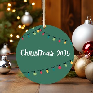 A green Christmas ornament with colorful lights and the text "Christmas 2025" is displayed on a wooden surface, surrounded by other Christmas ornaments and a blurred background of a Christmas tree.