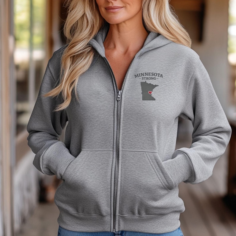 K&ouml;nnte beinhalten: Ein grauer Kapuzenpullover mit Rei&szlig;verschluss, Kapuze und Taschen. Der Hoodie tr&auml;gt den Text "MINNESOTA STRONG" &uuml;ber einer grauen Umrisslinie des Staates mit einem roten Herz. Der Rei&szlig;verschluss ist silberfarben.