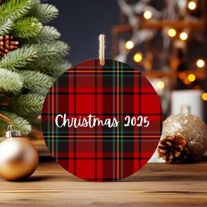 A Christmas ornament with a red and black plaid pattern, featuring the text "Christmas 2025" on it, is displayed on a wooden surface. The ornament is surrounded by other Christmas decorations, including a Christmas tree, ornaments, and a glittery ball.