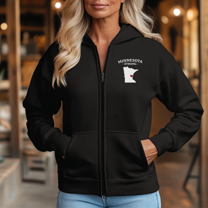 K&ouml;nnte beinhalten: Schwarzer Kapuzenpullover mit Rei&szlig;verschluss, Kapuze und Vordertaschen. Auf der linken Brust befindet sich der Text "MINNESOTA STRONG" &uuml;ber einer wei&szlig;en Umrisslinie des Bundesstaates Minnesota mit einem roten Herz. Der Hoodie hat einen durchgehenden Rei&szlig;verschluss.