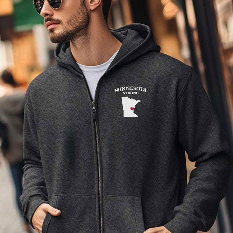 K&ouml;nnte beinhalten: Anthrazitfarbener Kapuzenpullover mit Rei&szlig;verschluss, der den Text "MINNESOTA STRONG" und eine wei&szlig;e Umrisslinie des Bundesstaates Minnesota mit einem roten Herzen zeigt. Der Hoodie hat eine Kapuze und einen Frontrei&szlig;verschluss.