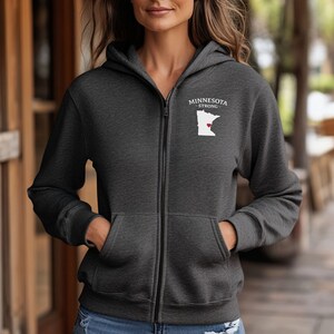 K&ouml;nnte beinhalten: Anthrazitfarbener Kapuzenpullover mit Rei&szlig;verschluss, mit einer wei&szlig;en Umrisszeichnung von Minnesota und einem roten Herz, sowie dem Text "MINNESOTA - STRONG" dar&uuml;ber. Der Hoodie hat einen durchgehenden Rei&szlig;verschluss, Vordertaschen und eine Kapuze.
