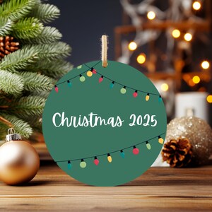A green circular ornament with the text "Christmas 2025" written on it, surrounded by Christmas lights and ornaments, with a blurred background of a Christmas tree and other holiday decorations.