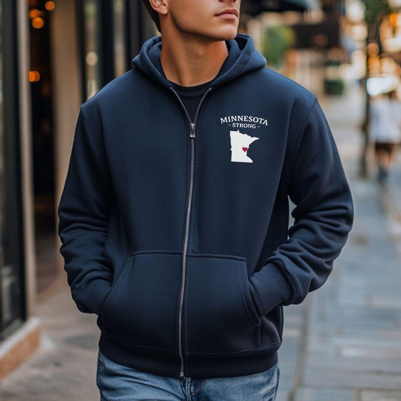 K&ouml;nnte beinhalten: Marineblauer Kapuzenpullover mit Rei&szlig;verschluss und Vordertaschen. Der Hoodie tr&auml;gt den Text "MINNESOTA STRONG" &uuml;ber einer wei&szlig;en Umrisslinie des Bundesstaates mit einem roten Herz. Der Rei&szlig;verschluss verl&auml;uft mittig.