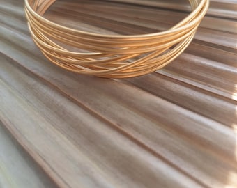 May include: A gold-colored cuff bracelet with a woven design. The bracelet is made of multiple thin, parallel strands that curve around to form a circular shape. The bracelet is approximately 2.5 inches in diameter.