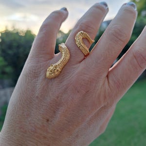 May include: A gold-coloured snake ring wraps around a finger. The ring has a textured design, with the snake's head and tail meeting on the finger. The ring is a unique piece of jewellery.