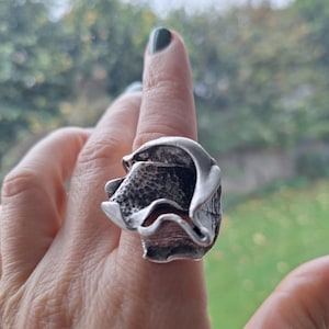 May include: A silver ring with an abstract, textured design. The ring has a layered, organic shape with a hammered texture on the inner surface. The ring is worn on a finger, showcasing its unique, sculptural form.