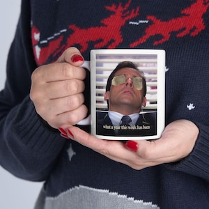 Puede incluir: Taza de cerámica blanca con una foto de un hombre con bolsitas de té sobre los ojos y el texto "what a year this week has been". La taza la sostiene una persona que lleva un jersey azul con un estampado de renos rojos.