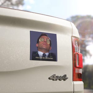 May include: A square vinyl sticker featuring a man wearing gold-rimmed glasses with his head tilted back. The image includes the text 'what a year this week has been' in white lettering at the bottom. The sticker is applied to a light-colored vehicle.