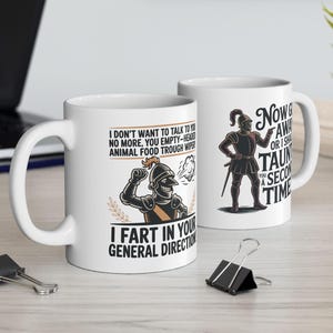 May include: Two white ceramic mugs with humorous text and illustrations. One mug features a knight illustration and the phrase "I FART IN YOUR GENERAL DIRECTION." The other mug has a knight illustration and the phrase "Now Away or I Shall Taunt You a Second Time."