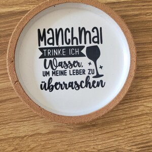 Może przedstawiać: Okrągła podstawka korkowa z białym środkiem zawierającym czarny napis: Manchmal TRINKE ICH Wasser, UM MEINE LEBER ZU überraschen, któremu towarzyszy czarna sylwetka kieliszka do wina oraz dwie małe ikony gwiazdek na białej powierzchni.