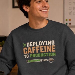 May include: A dark charcoal gray crewneck sweatshirt featuring a graphic print with the text "DEPLOYING CAFFEINE TO PRODUCTION" in white, tan, and green. Below the text is a green loading bar at 100% and a small coffee cup icon with steam.