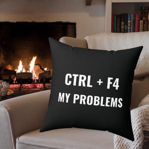 May include: Black square throw pillow with the text "CTRL + F4 MY PROBLEMS" in white. The pillow is on a beige armchair in front of a fireplace with a burning fire.