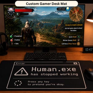 May include: Black desk mat with the text "Human.exe has stopped working" and a warning symbol. The mat is under a keyboard and a computer monitor displaying a video game scene. The text "Custom Gamer Desk Mat" is at the top.