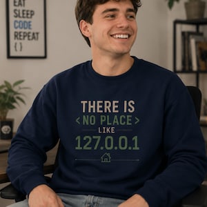 May include: A navy blue crewneck sweatshirt featuring the text "THERE IS <NO PLACE> LIKE 127.0.0.1" printed in light beige and muted green. The design includes a small graphic of a house icon centered below the IP address text.