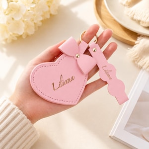 Peut inclure: Un porte-clés rose clair en forme de cœur avec un nœud décoratif sur le dessus et une lanière assortie à bords festonnés. Le cœur et la lanière portent le nom Liliana gravé en écriture cursive. L'objet comprend un anneau en métal doré.